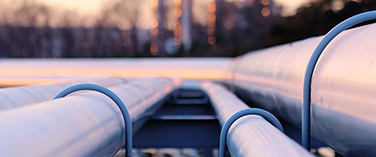 Close-up of a series of industrial steel pipelines for oil and natural gas transport, set against a blurred industrial background at sunset.